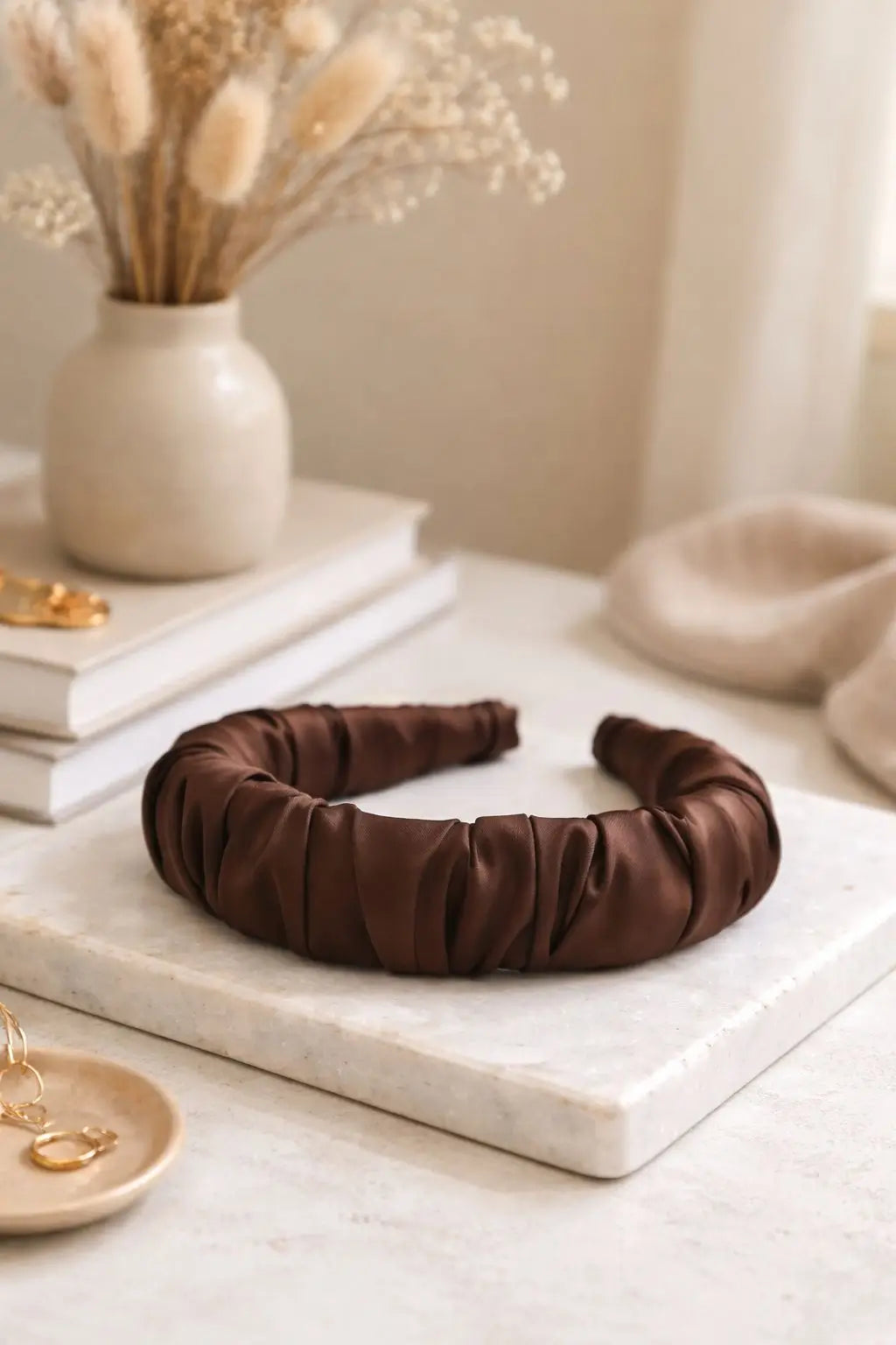 A dark brown ruched satin headband displayed on a white marble tray, styled with dried flowers in a ceramic vase and gold jewelry on a light marble surface.
