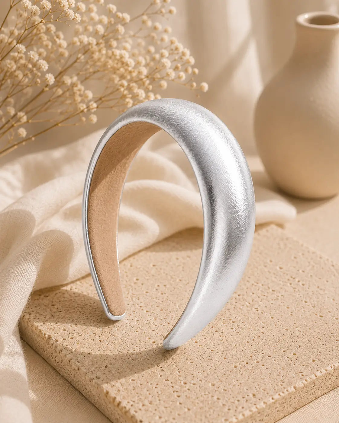 A wide, curved silver metal headband with a textured, brushed finish and a tan interior lining, displayed on a beige textured stone block with neutral-toned fabric and dried flowers in the background.