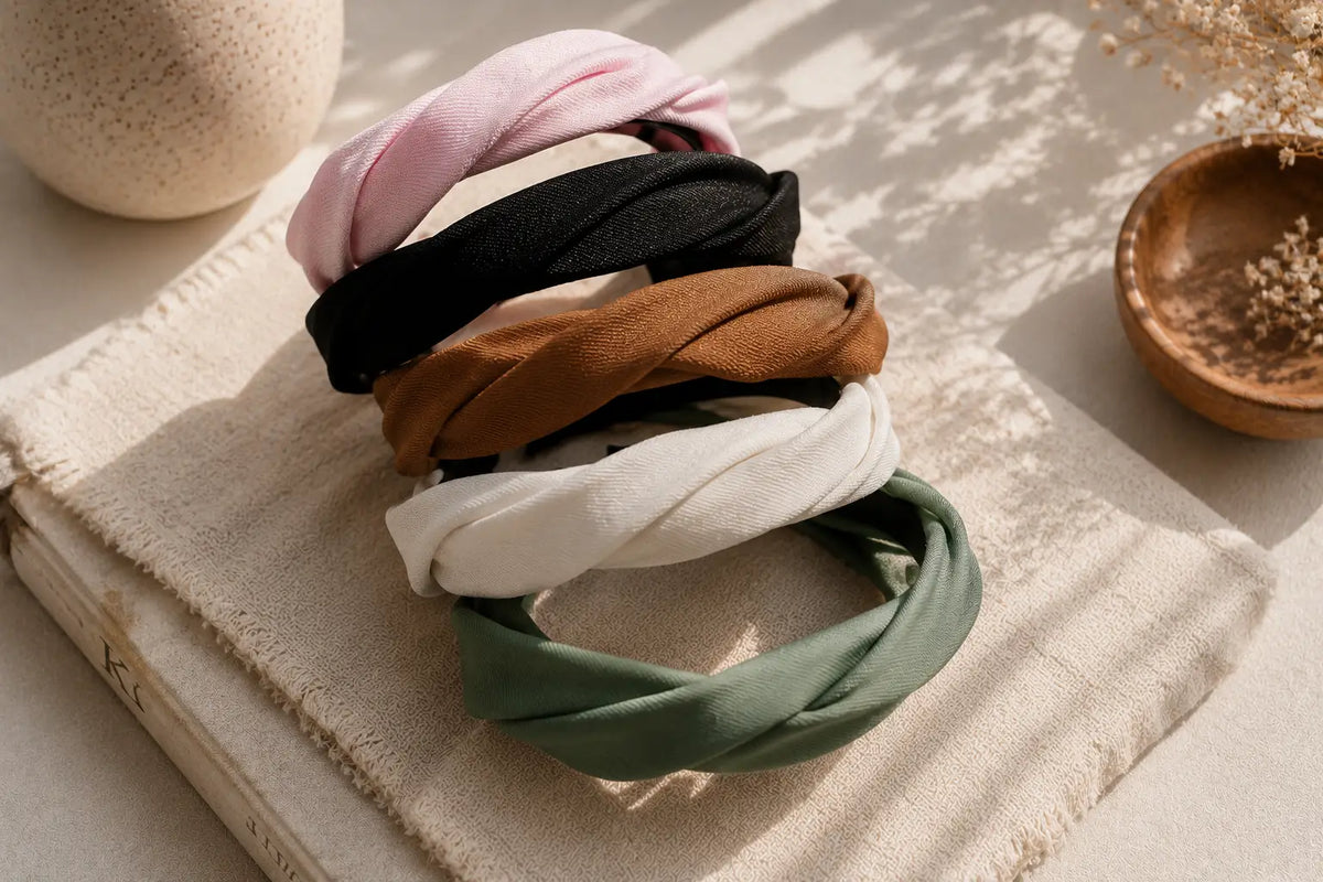 Five twisted fabric headbands in pink, black, brown, white, and sage green are stacked on a textured cream fabric surface with a book and dried floral decor in the background.