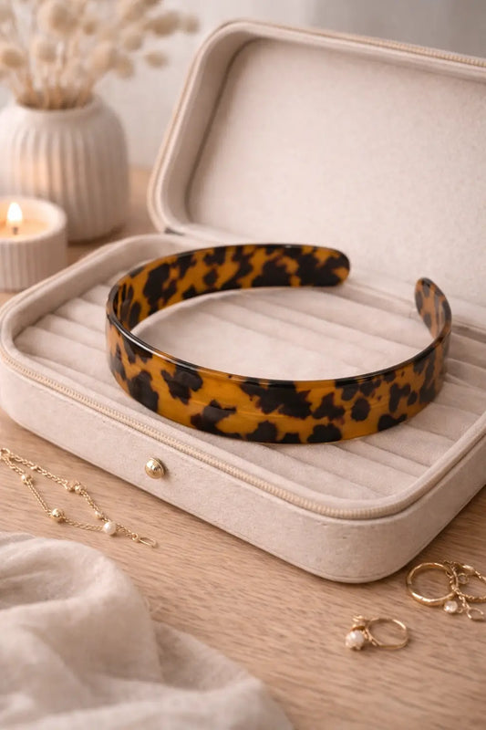 A tortoiseshell acetate headband with a yellow-orange and black leopard pattern, displayed inside an open beige velvet jewelry box on a wooden table with styled props.