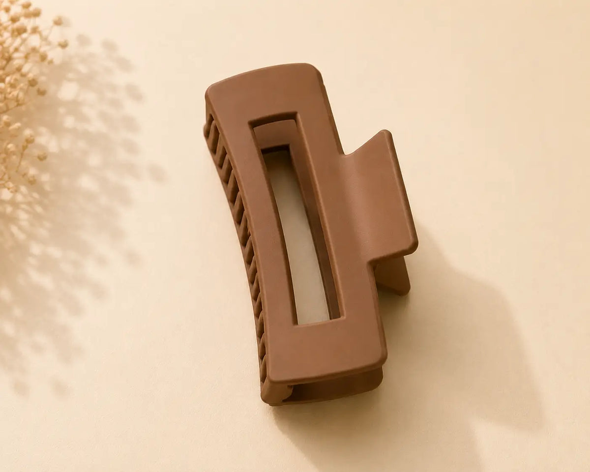 A single large hair claw clip in a matte warm brown color, photographed on a light beige background with soft decorative dried flowers in the upper left corner.