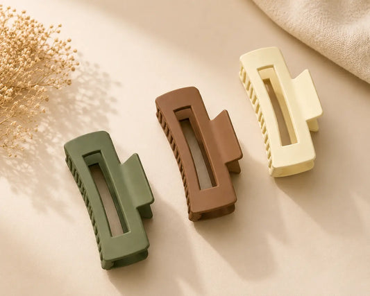 Three hair claw clips in sage green, medium brown, and cream are arranged diagonally on a warm beige surface with soft shadows, dried flowers, and a textured fabric in the background.