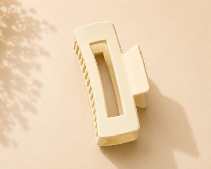 A cream-colored rectangular hair claw clip with a center cutout, shown on a warm beige background with soft leaf shadows.