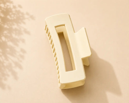 A cream-colored rectangular hair claw clip with a center cutout, shown on a warm beige background with soft leaf shadows.