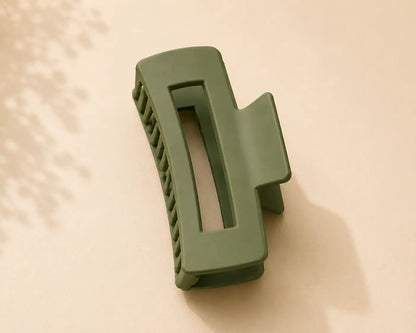 An olive green hair claw clip with a rectangular center and ridged outer edges, resting on a warm beige surface with soft leaf shadows.