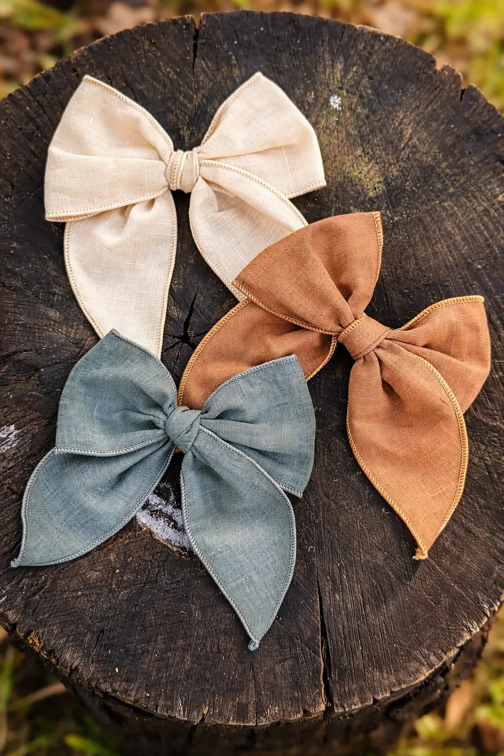 Three fabric hair bows in cream, tan, and dusty blue-green are arranged on a dark wood tree stump outdoors.