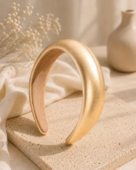 A wide, curved gold headband with a matte metallic finish, displayed on a textured beige stone slab with draped cream fabric and dried flowers in the background.