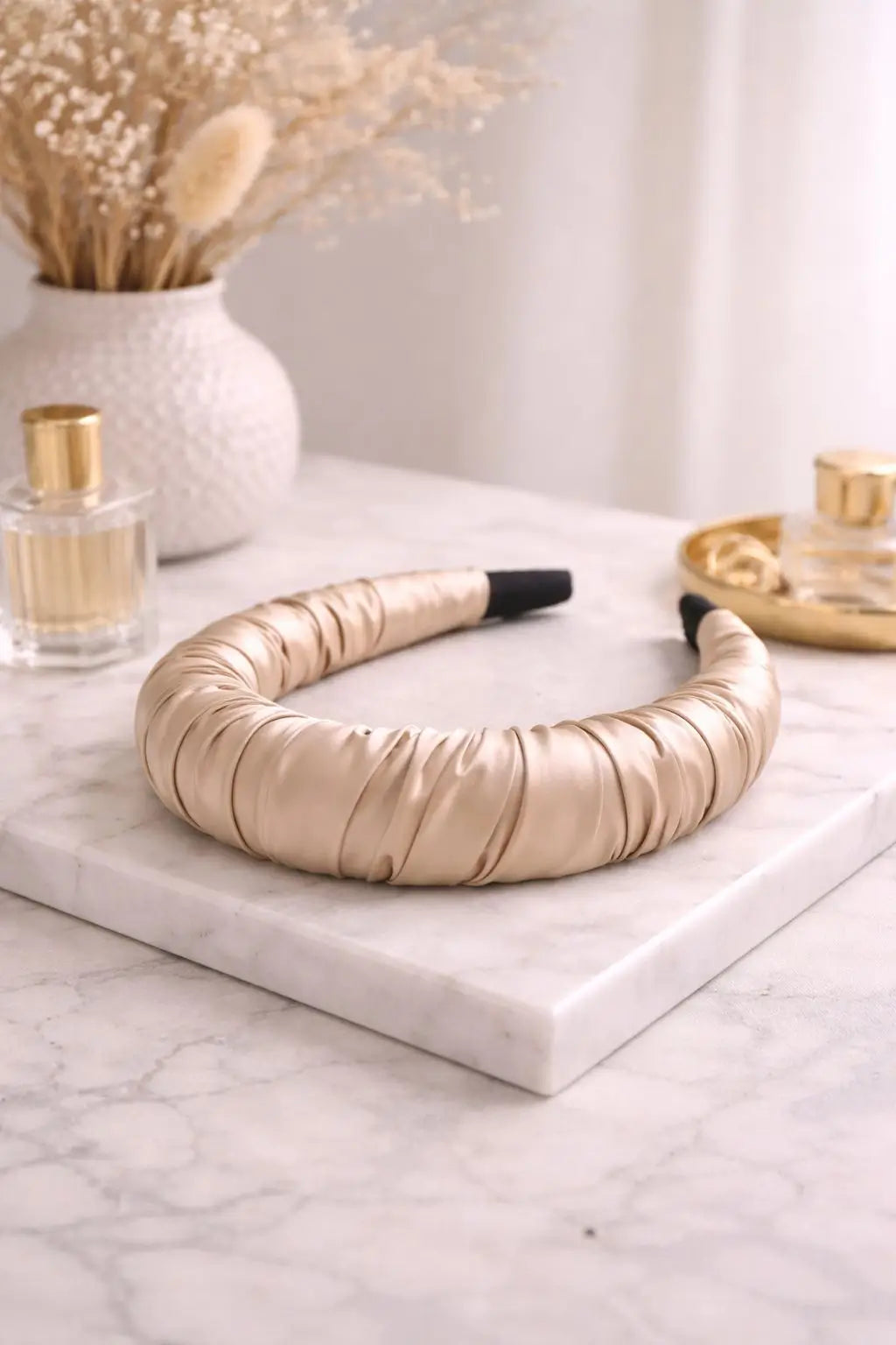A padded champagne gold satin headband with black tips, displayed on a white marble tray with decorative perfume bottles and dried flowers in the background.
