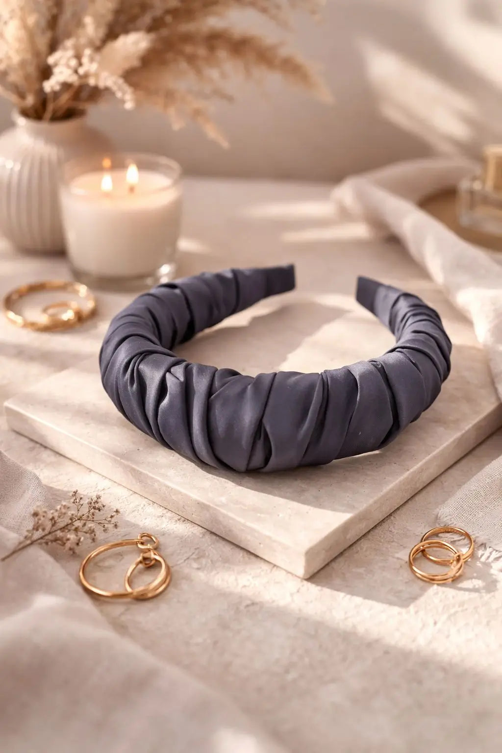 A wide, gathered fabric headband in a muted slate blue-gray color, styled on a light stone block with gold hoop earrings, a lit candle, and dried flowers.