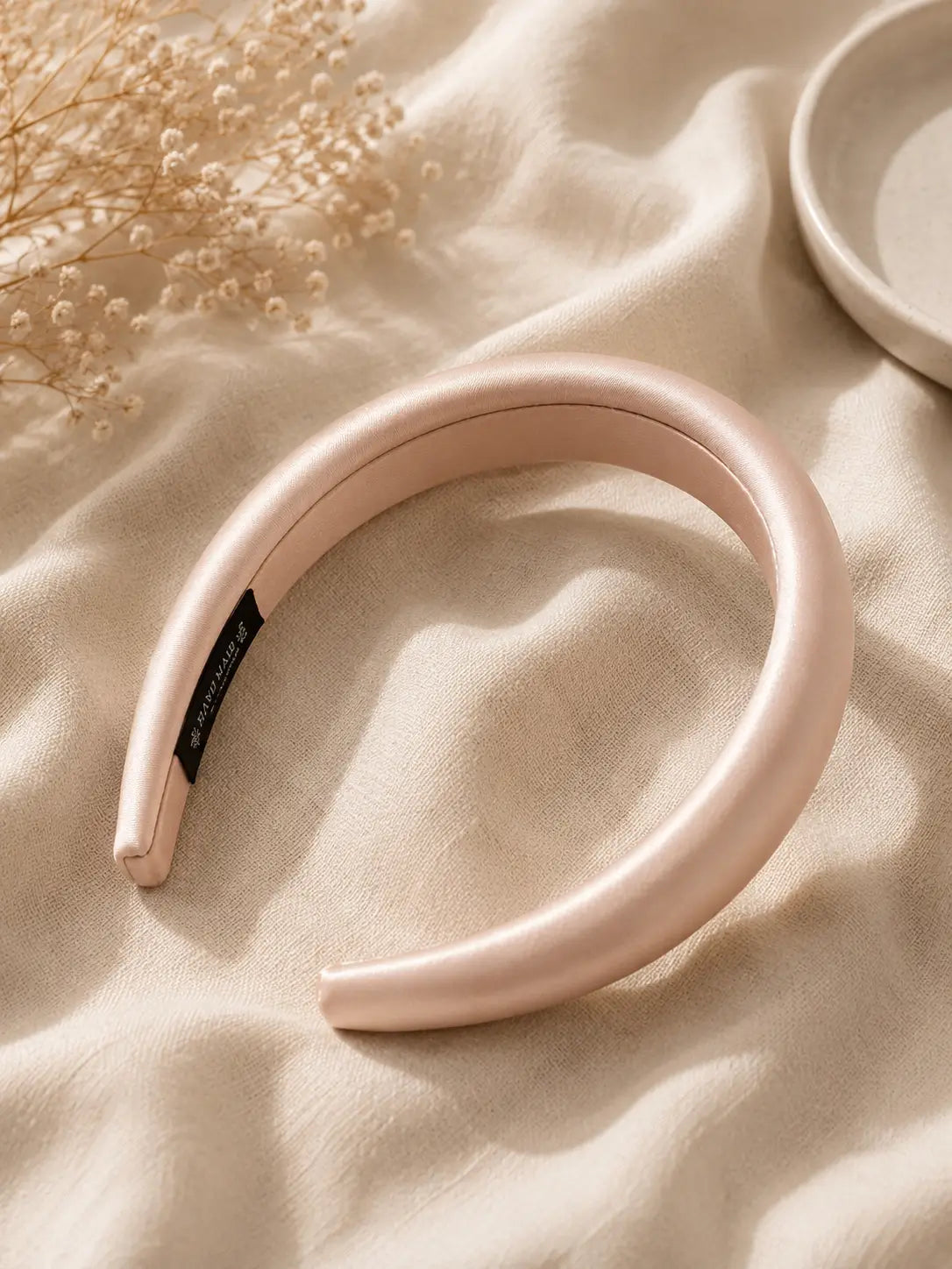 A wide, padded headband in a soft blush pink color, styled on a draped beige fabric with dried baby's breath flowers and a ceramic dish.
