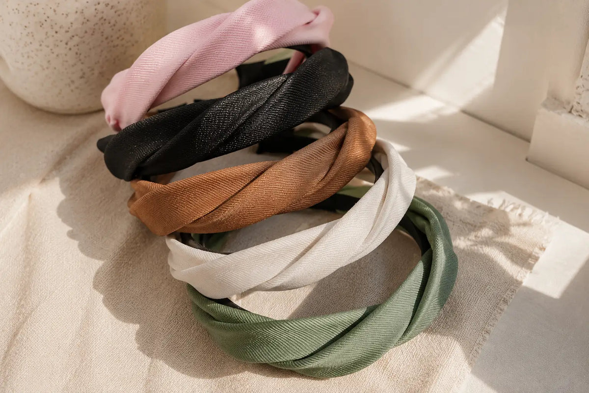 Five twisted fabric headbands in pink, black, tan, off-white, and sage green, stacked on a textured beige fabric surface with warm natural sunlight and shadows.