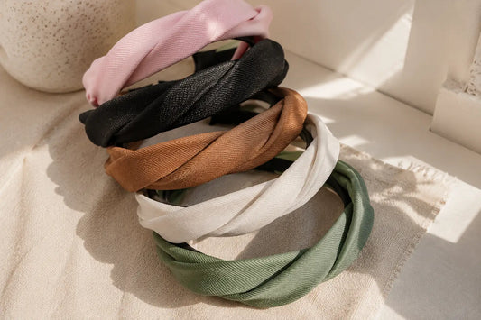 Five twisted fabric headbands in pink, black, tan, off-white, and sage green, stacked on a textured beige fabric surface with warm natural sunlight and shadows.
