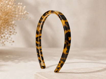 A glossy tortoiseshell hair headband with a yellow, black, and brown mottled pattern, standing upright on a light beige pedestal against a warm beige background.