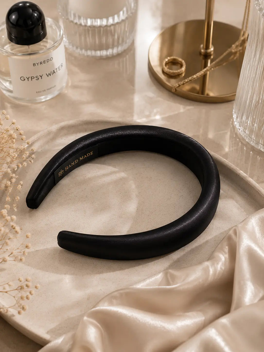 A wide, padded black headband with a matte finish, displayed on a beige satin cloth with a tray, silk scarf, and decorative props including a perfume bottle and gold jewelry stand.